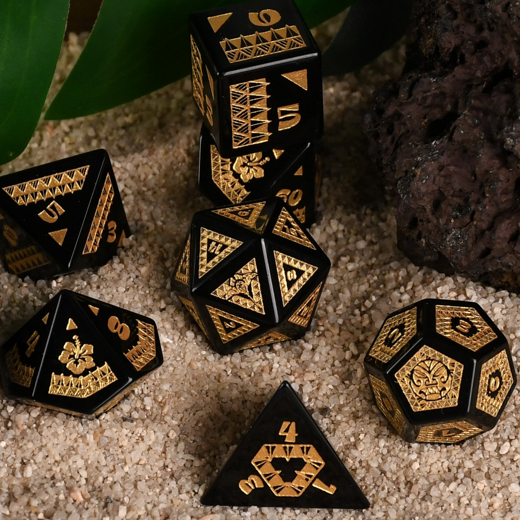 A set of black obsidian stone dice with gold engravings, displayed on a sandy surface with a green plant in the background.