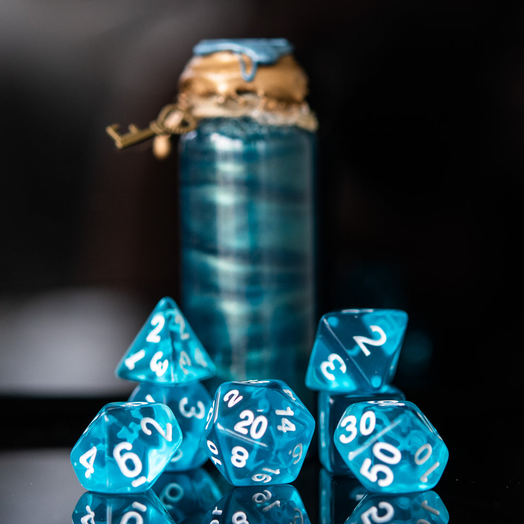 A set of blue acrylic dice with engraved numbers, displayed in front of a small bottle-shaped container with a cork. The dice include a D4, D6, D8, two D10s, D12, and D20.