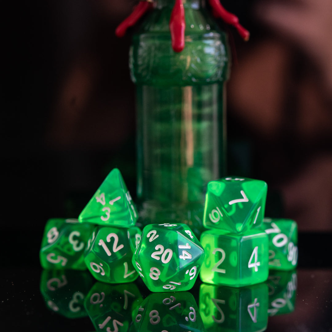 A set of green acrylic dice with engraved numbers, displayed in front of a small bottle with a red stopper. The dice include a D4, D6, D8, two D10s, D12, and D20, arranged on a reflective surface.