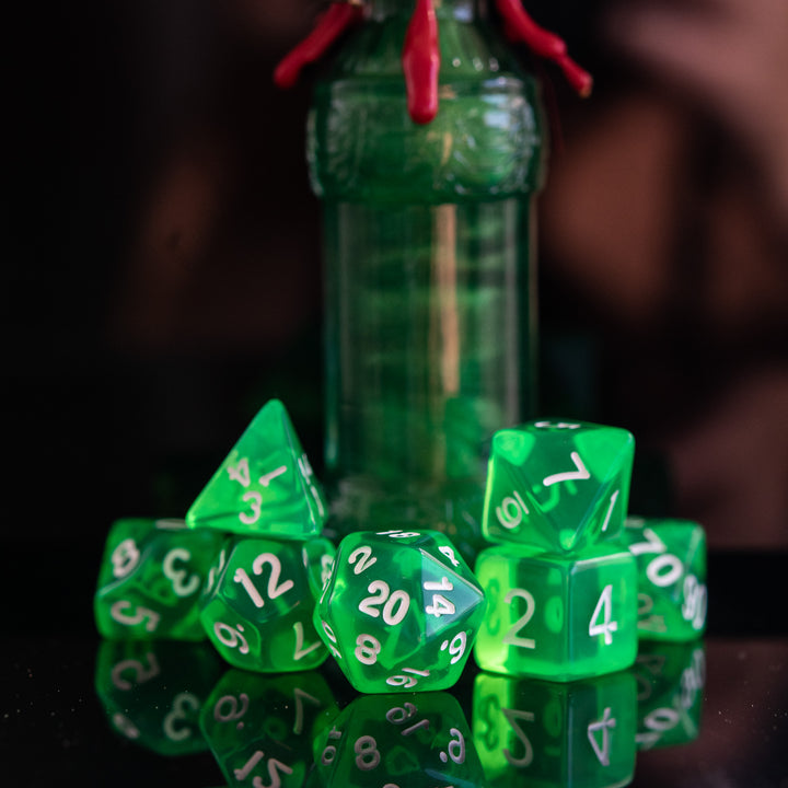 A set of green acrylic dice with engraved numbers, displayed in front of a small bottle with a red stopper. The dice include a D4, D6, D8, two D10s, D12, and D20, arranged on a reflective surface.