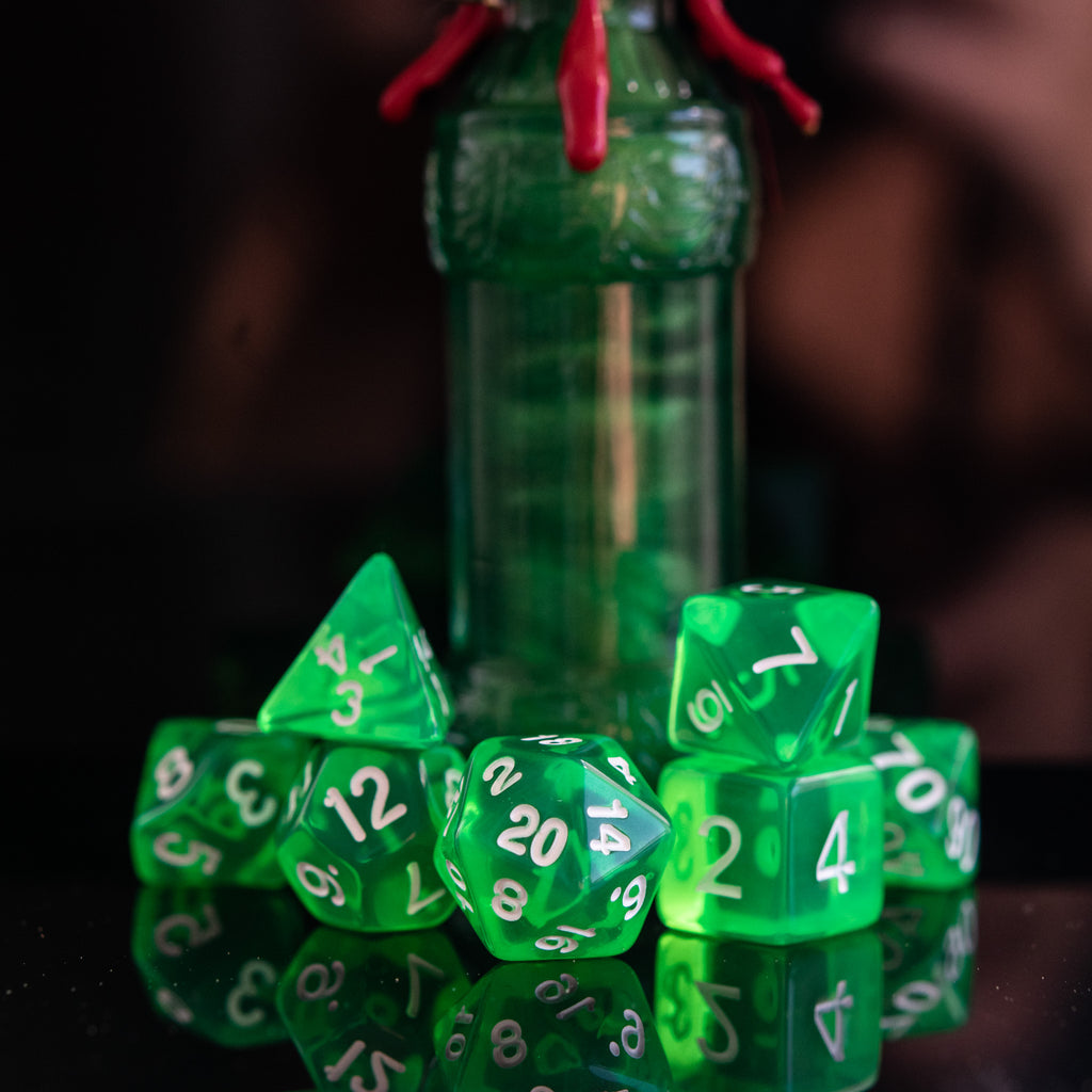 A set of green acrylic dice with engraved numbers, displayed in front of a small bottle with a red stopper. The dice include a D4, D6, D8, two D10s, D12, and D20, arranged on a reflective surface.