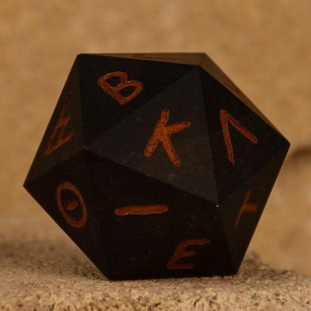 A 30mm black serpentine stone d20 dice with ancient Greek letters inscribed on each face, positioned on a textured surface.