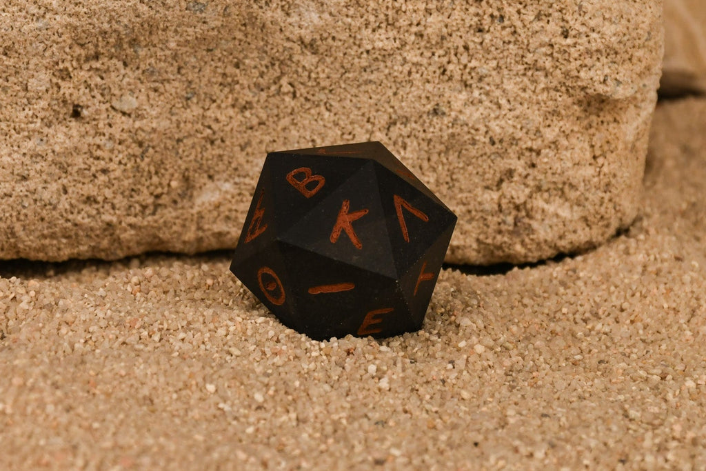 A 30mm black serpentine stone d20 dice with ancient Greek letters inscribed on each face, positioned on a textured surface.
