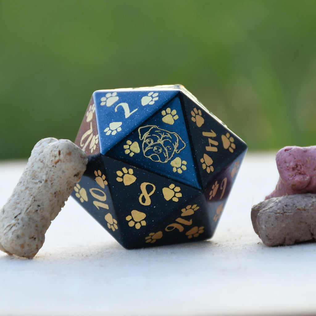 A hand-carved blue sandstone dice with puppy paw designs, placed next to a bone-shaped object, against a blurred green background.