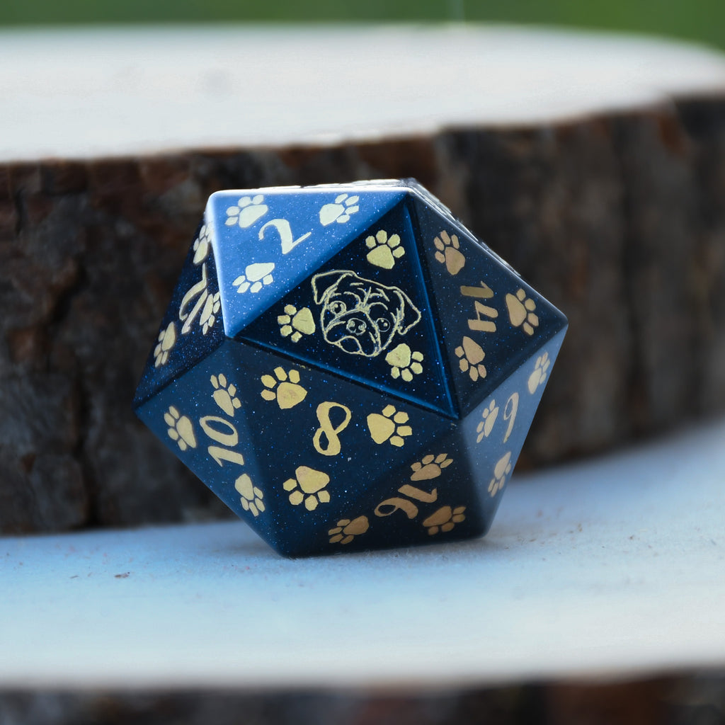 A hand-carved blue sandstone dice with puppy paw designs, placed next to a bone-shaped object, against a blurred green background.