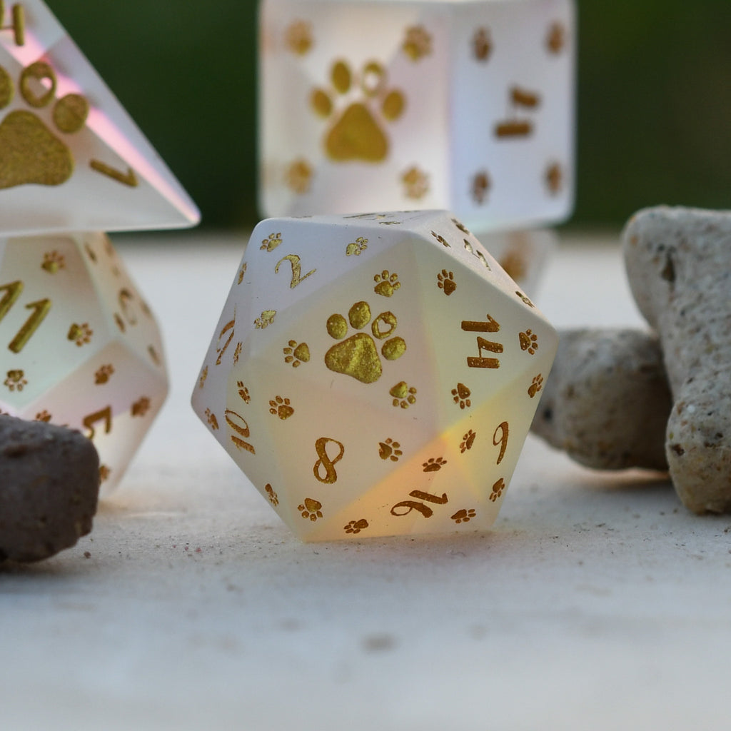 A set of frosted glass dice with gold paw prints and numbers, including various shapes typical for role-playing games like D&D.