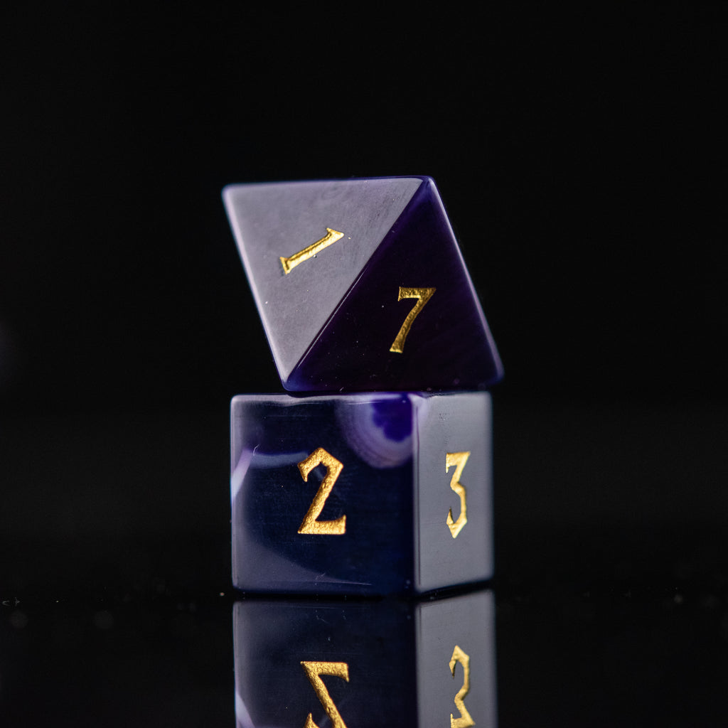 A set of purple agate stone dice with engraved numbers, displayed on a reflective surface.