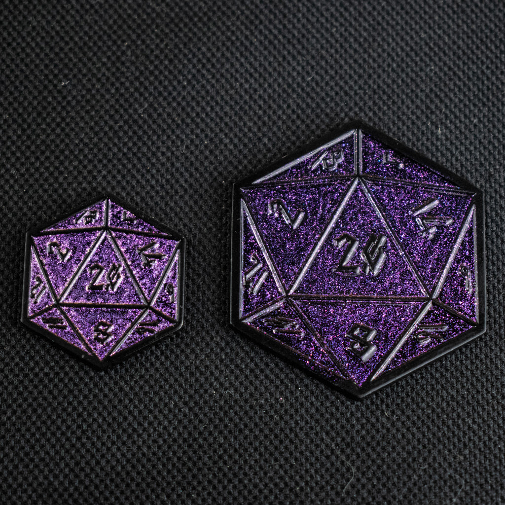 Two purple glittering D20 pins of different sizes placed on a textured black surface.