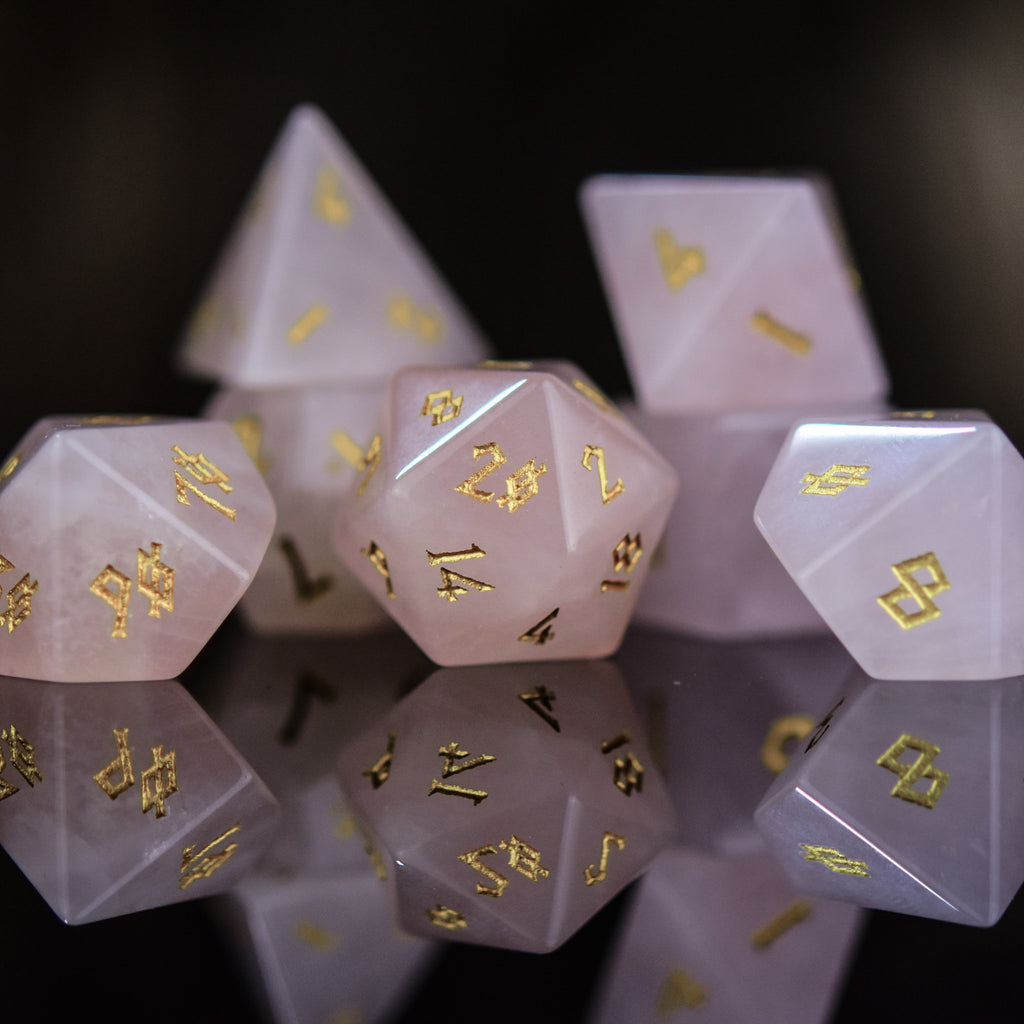 A collection of pink rose quartz stone dice with engraved numbers, displayed on a reflective surface.