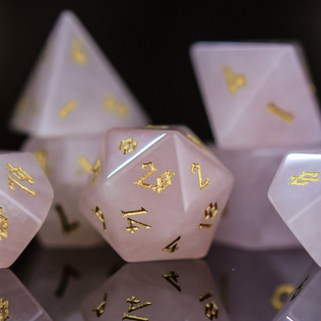 A collection of pink rose quartz stone dice with engraved numbers, displayed on a reflective surface.