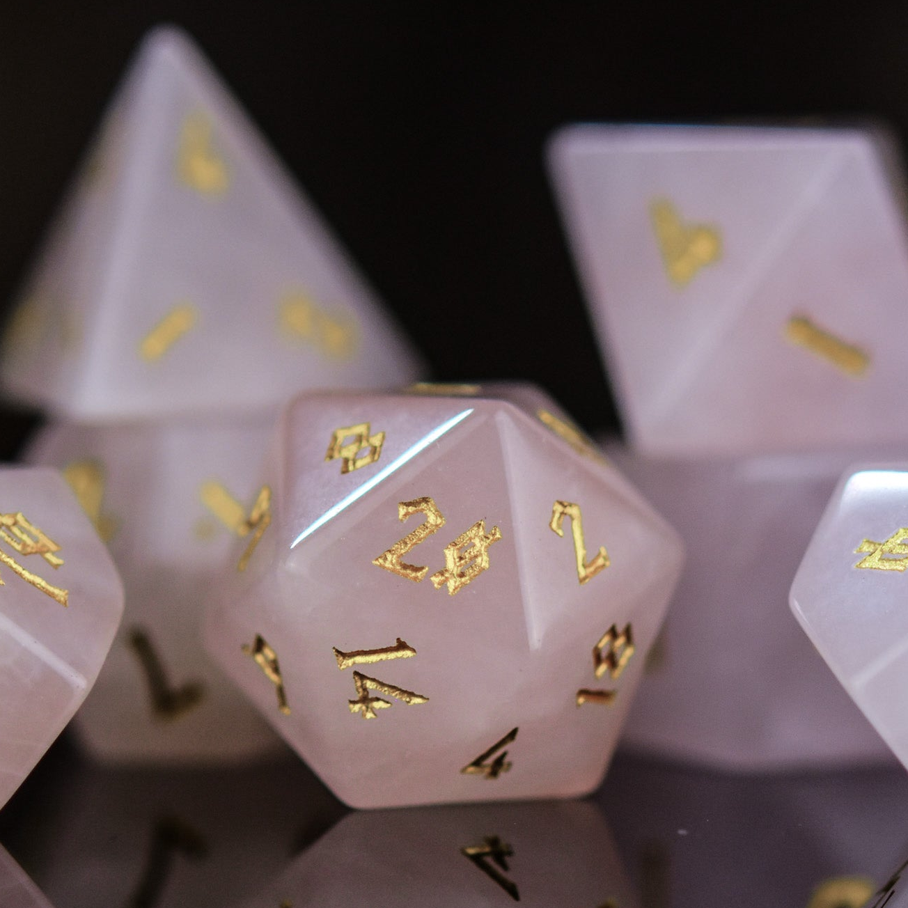A collection of pink rose quartz stone dice with engraved numbers, displayed on a reflective surface.