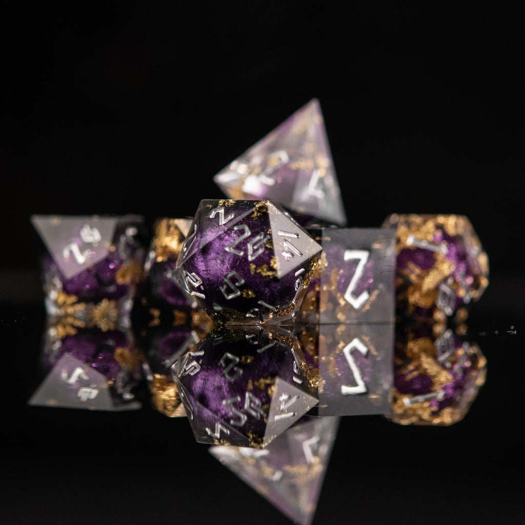 A set of hand-crafted, resin dice with liquid core, featuring purple and gold colors with engraved numbers. The dice are displayed on a reflective surface.