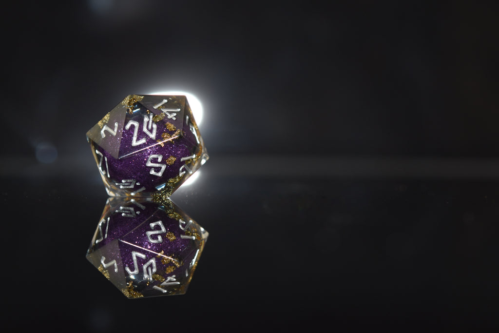 A set of hand-crafted, resin dice with liquid core, featuring purple and gold colors with engraved numbers. The dice are displayed on a reflective surface.