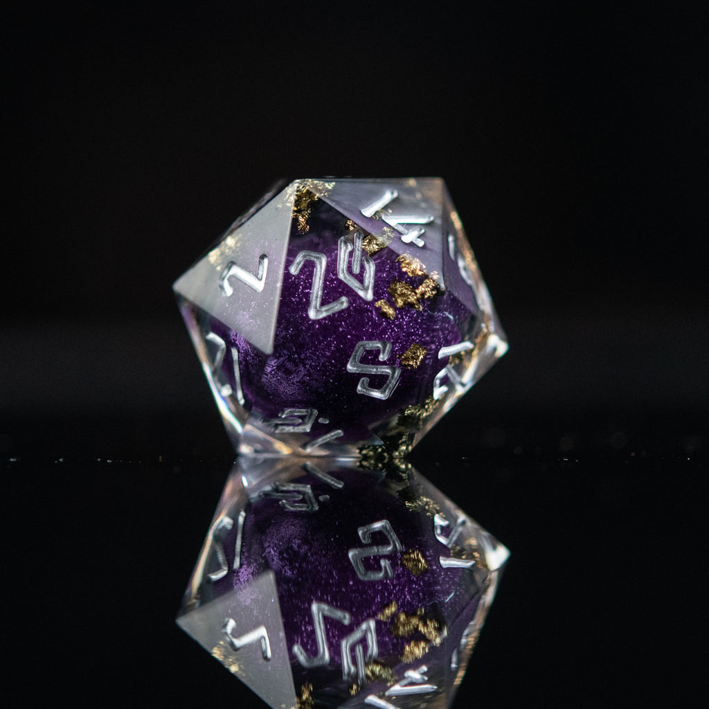 A set of hand-crafted, resin dice with liquid core, featuring purple and gold colors with engraved numbers. The dice are displayed on a reflective surface.