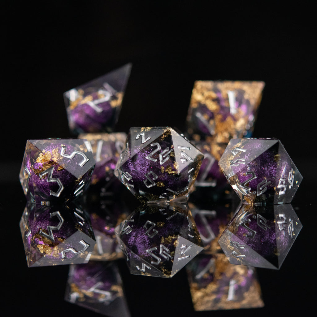 A set of hand-crafted, resin dice with liquid core, featuring purple and gold colors with engraved numbers. The dice are displayed on a reflective surface.