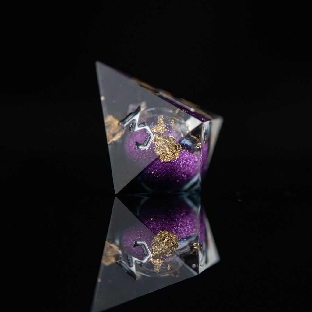 A set of hand-crafted, resin dice with liquid core, featuring purple and gold colors with engraved numbers. The dice are displayed on a reflective surface.
