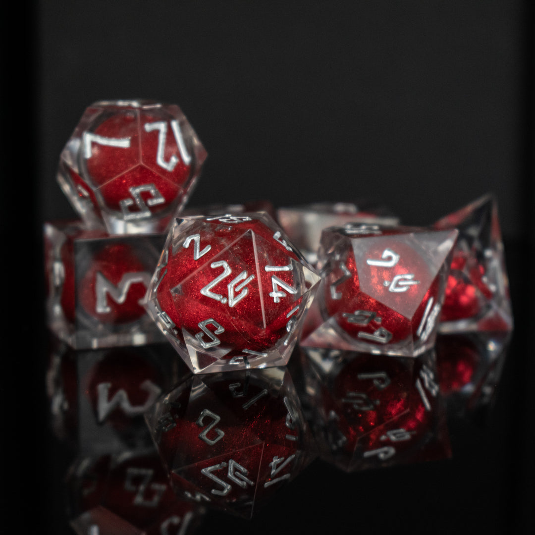 A set of clear resin dice with red liquid core and white hand-painted numbers, including various shapes typical for role-playing games like a D4, D6, D8, D10, D12, and D20.
