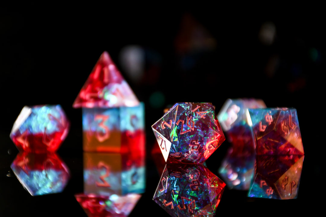A set of sharp-edged resin dnd dice in red and blue colors with hand-painted pink numbers and irridescent foil inside, reflecting on a surface.