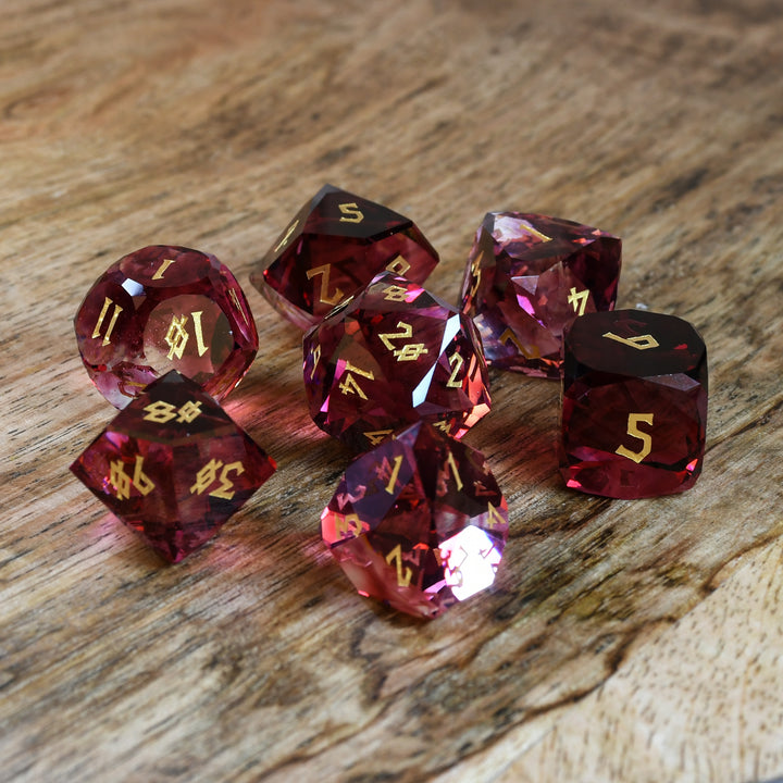 A set of multifaceted cut glass dnd dice with engraved numbers, displayed on a wooden surface. The dice are primarily red with gold text