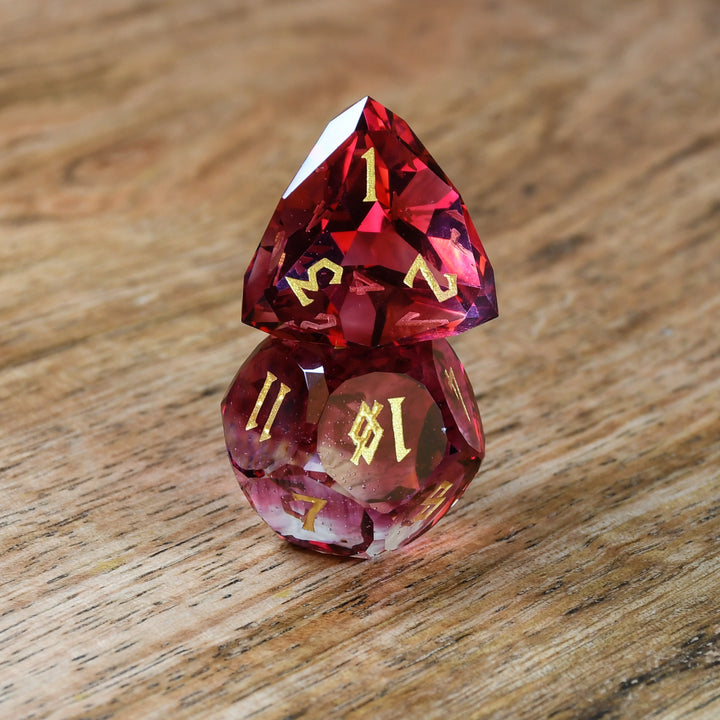 A set of multifaceted cut glass dnd dice with engraved numbers, displayed on a wooden surface. The dice are primarily red with gold text