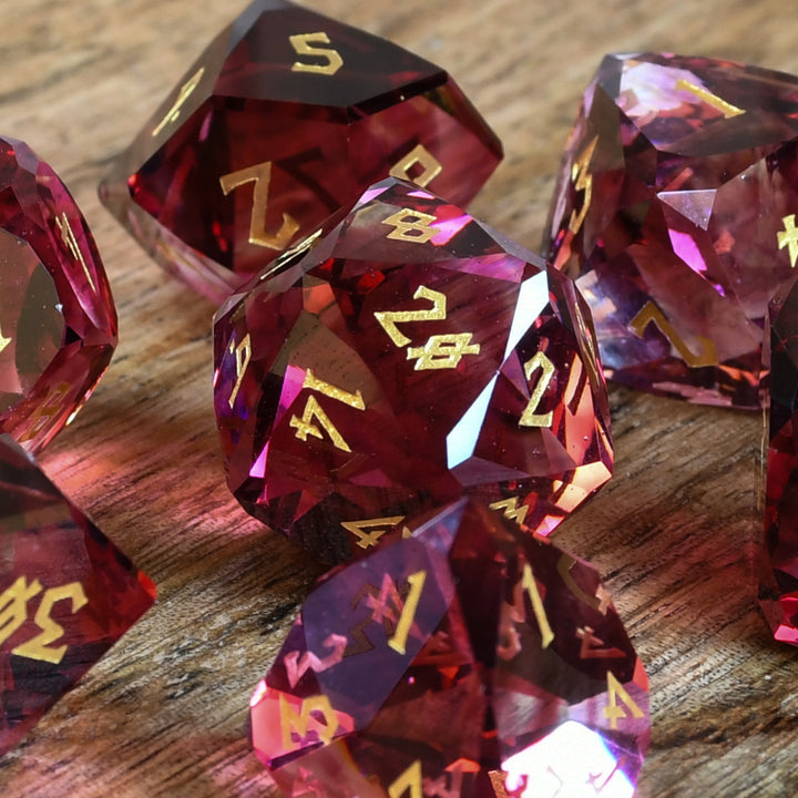 A set of multifaceted cut glass dnd dice with engraved numbers, displayed on a wooden surface. The dice are primarily red with gold text