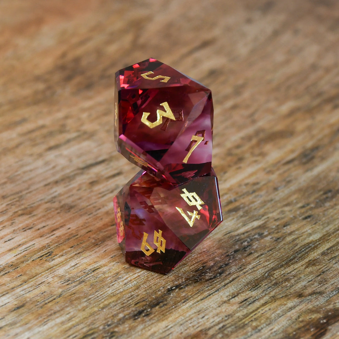 A set of multifaceted cut glass dnd dice with engraved numbers, displayed on a wooden surface. The dice are primarily red with gold text