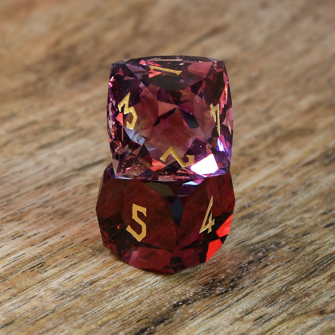 A set of multifaceted cut glass dnd dice with engraved numbers, displayed on a wooden surface. The dice are primarily red with gold text