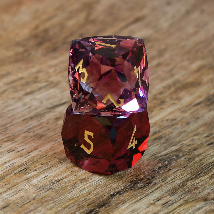 A set of multifaceted cut glass dnd dice with engraved numbers, displayed on a wooden surface. The dice are primarily red with gold text
