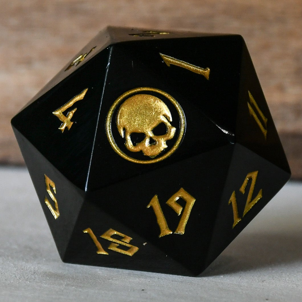 An oversized solid-metal black d20 spindown life counter die with gold numbers and an iconography designed for TCG gameplay.