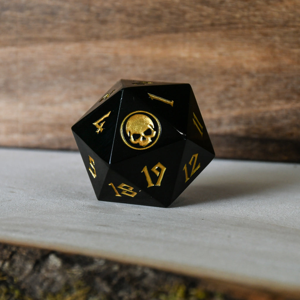 An oversized solid-metal black d20 spindown life counter die with gold numbers and an iconography designed for TCG gameplay.