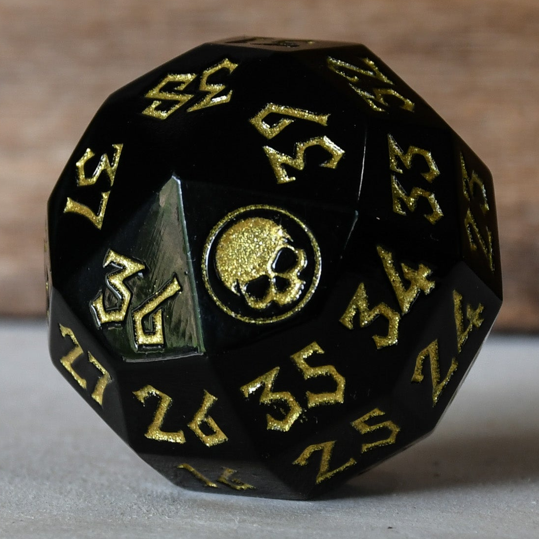 A solid-metal spindown life counter die used for trading card games, predominantly black with gold numbers and an icon of a skull on the face showing zero.