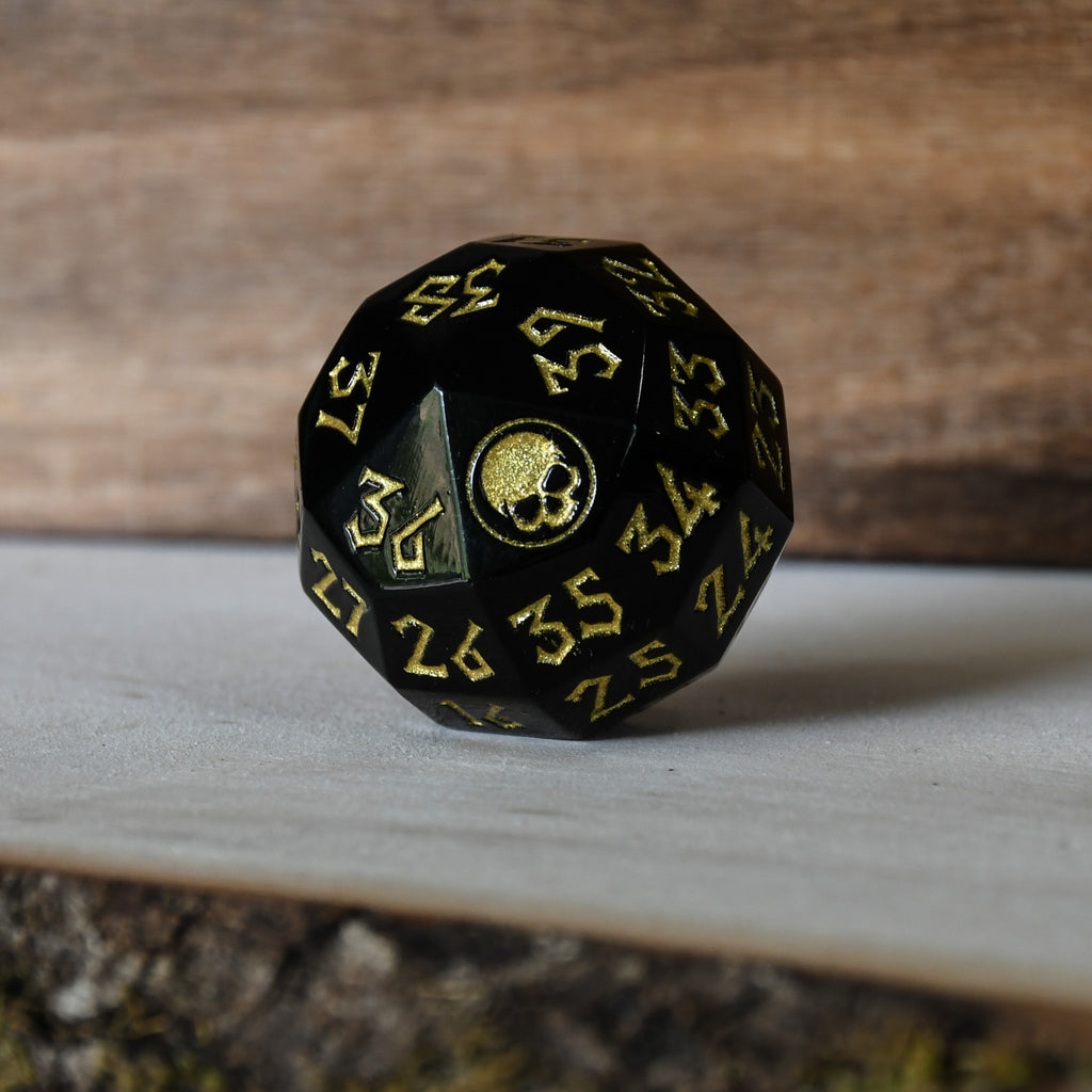 A solid-metal spindown life counter die used for trading card games, predominantly black with gold numbers and an icon of a skull on the face showing zero.