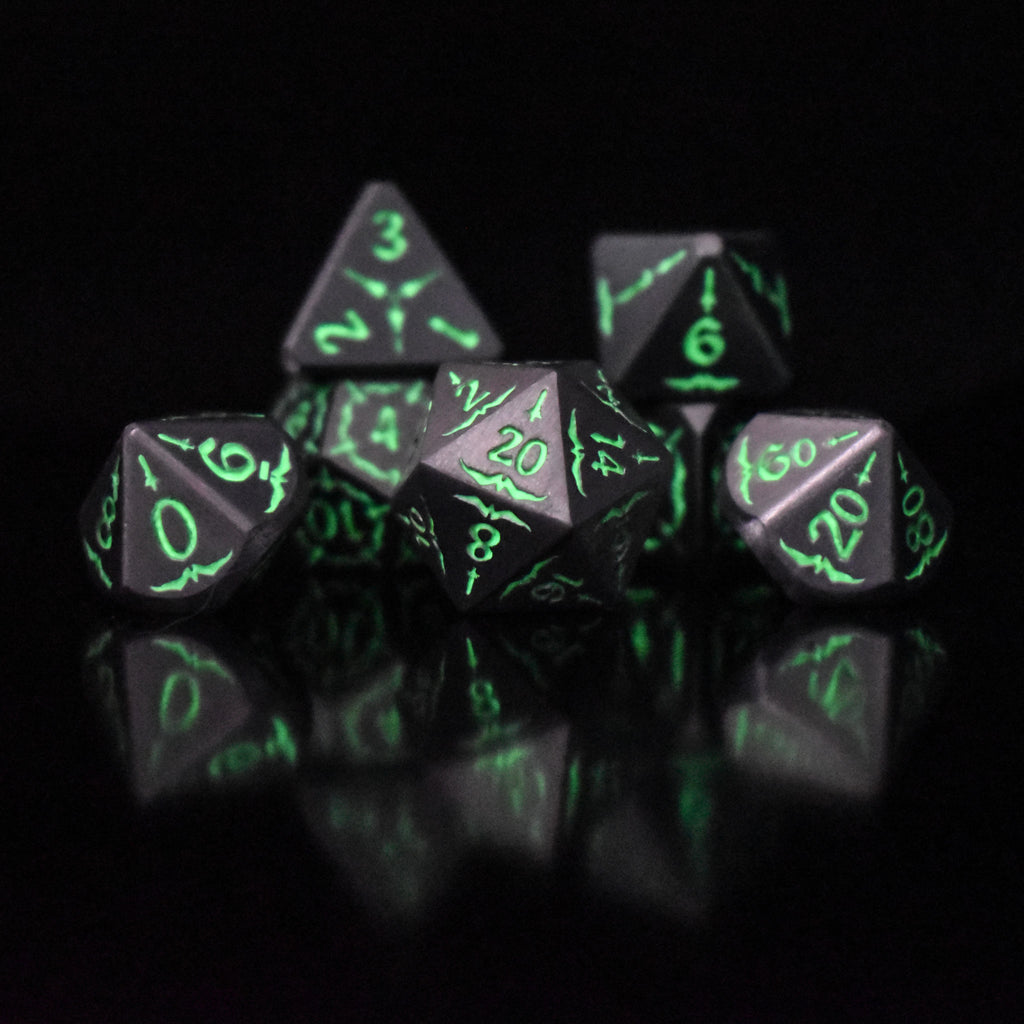 A set of glow-in-the-dark metal dnd dice with engraved numbers and swords, including various shapes such as a d4, d6, d8, d10, d12, and d20, displayed on a black background.