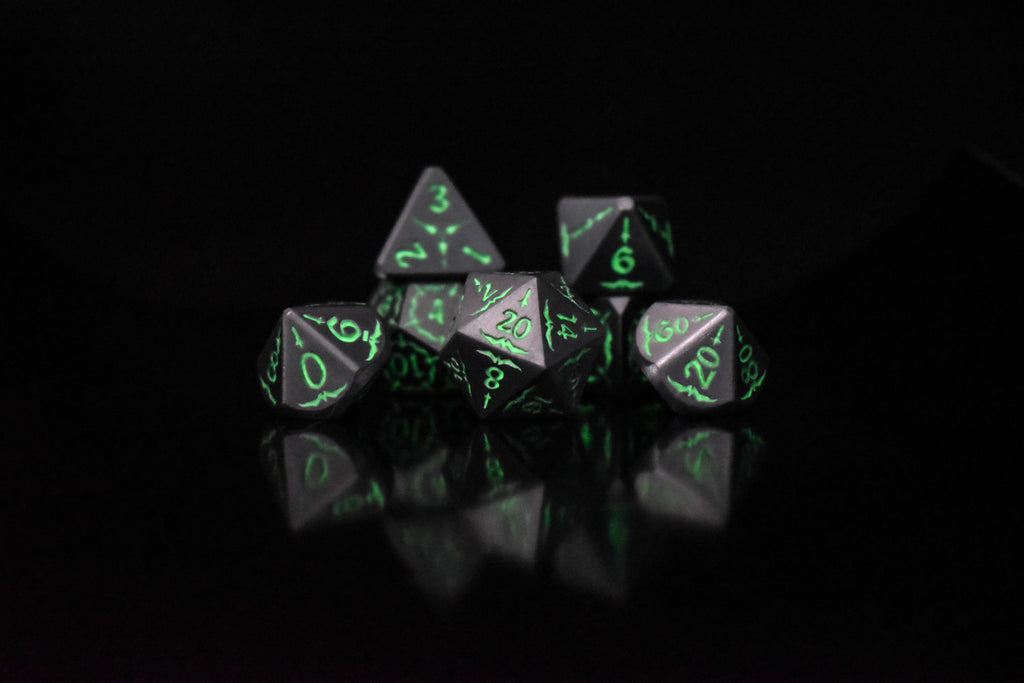 A set of glow-in-the-dark metal dnd dice with engraved numbers and swords, including various shapes such as a d4, d6, d8, d10, d12, and d20, displayed on a black background.