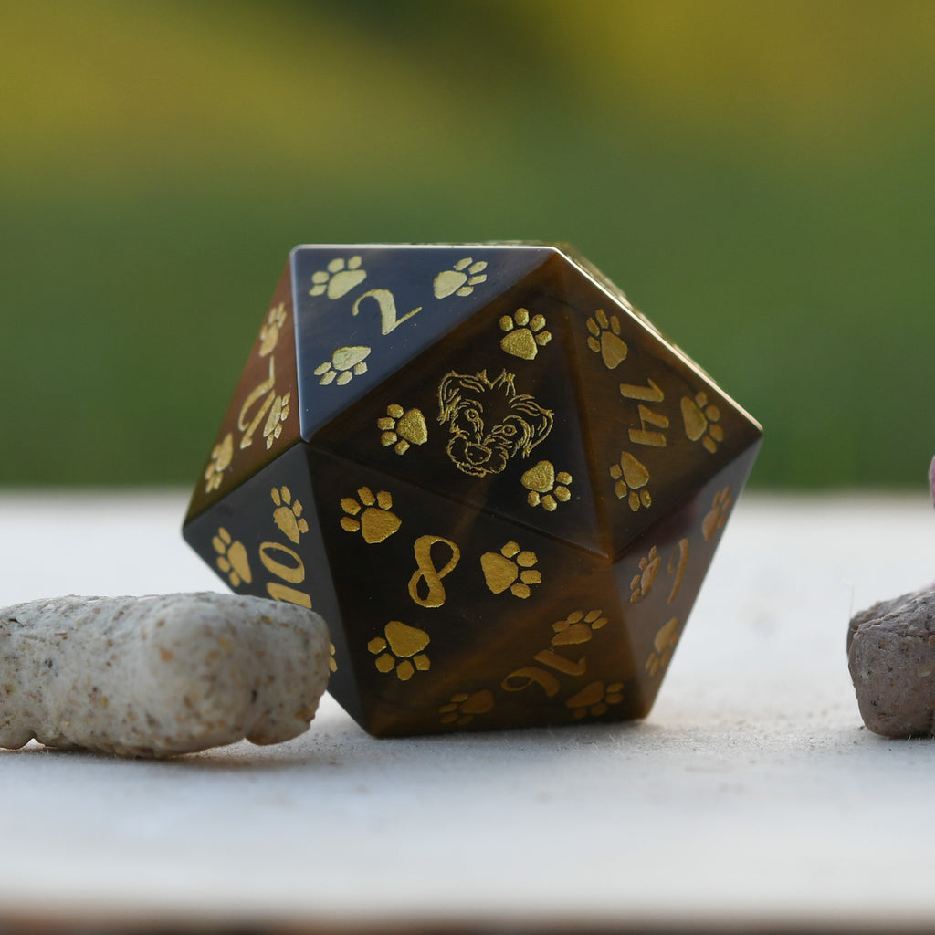 A Terrier Puppy Paws Tigers Eye 30mm D20 with puppy paw designs, engraved numbers and painted details, placed on a natural stone.