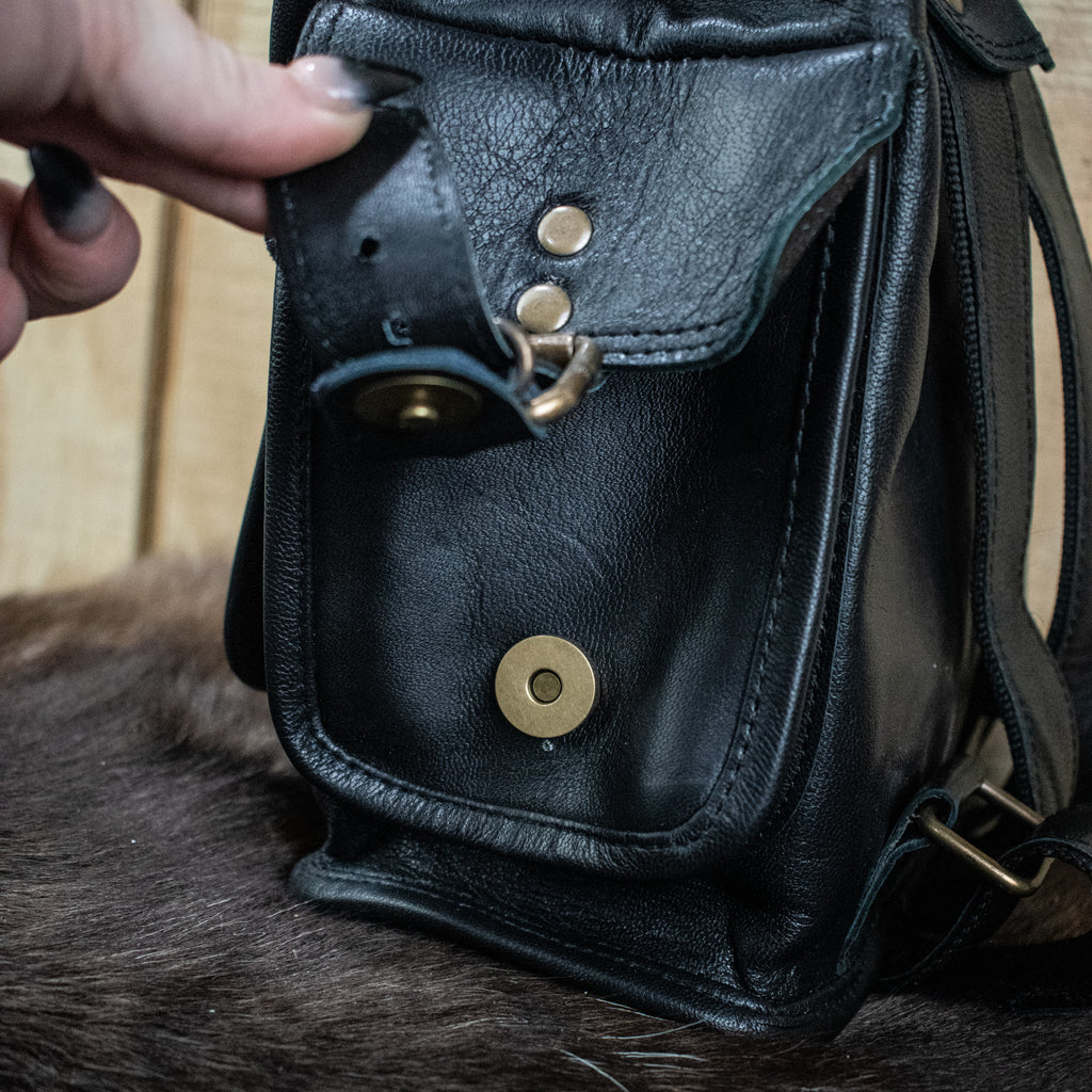 A black leather mini backpack with brass hardware, displayed against a wooden background.