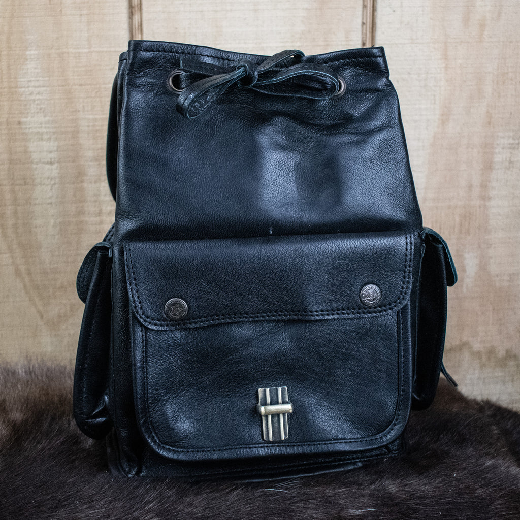 A black leather mini backpack with brass hardware, displayed against a wooden background.