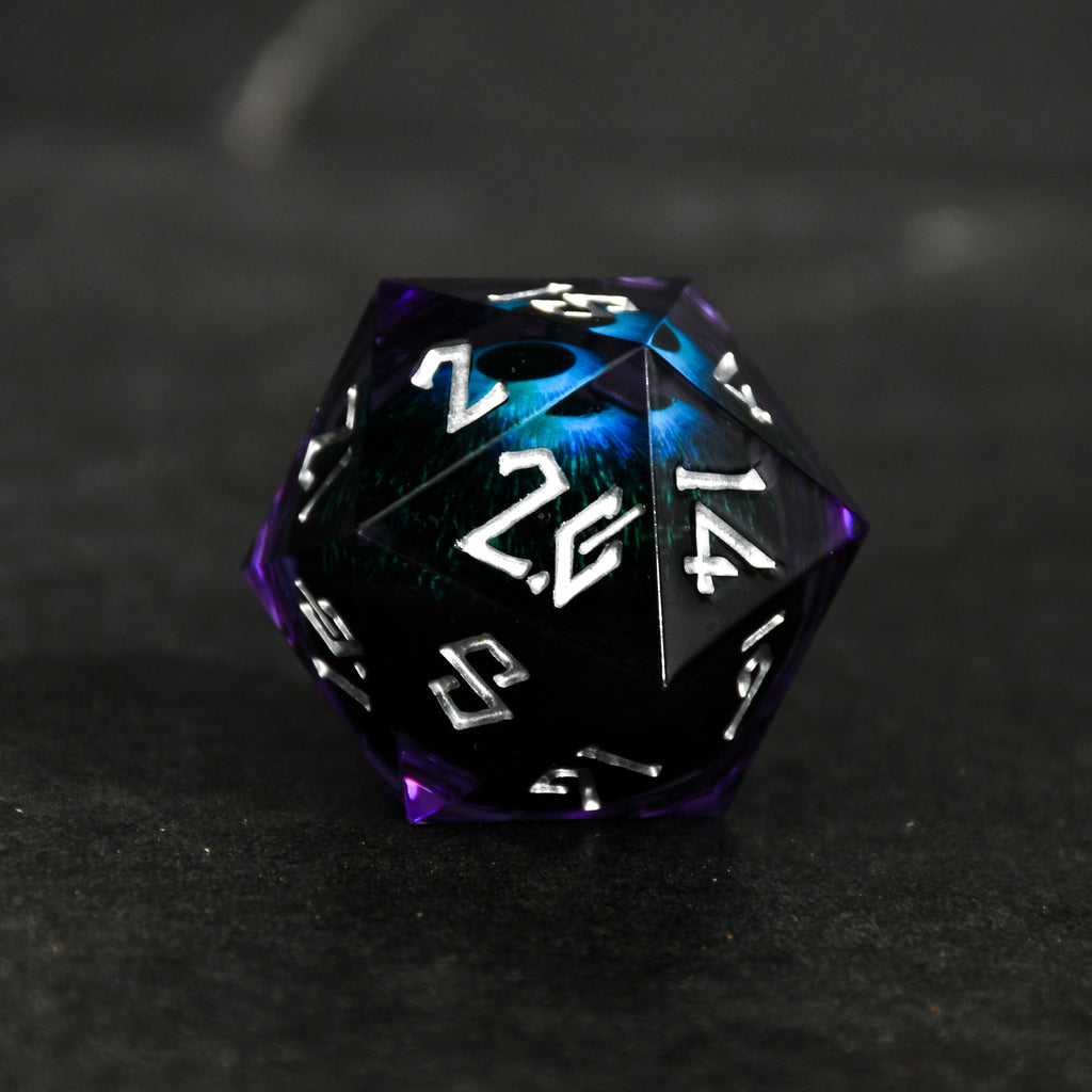 A large, multicolored resin dice with engraved numbers and a floating eye illustration inside, set against a textured grey background.