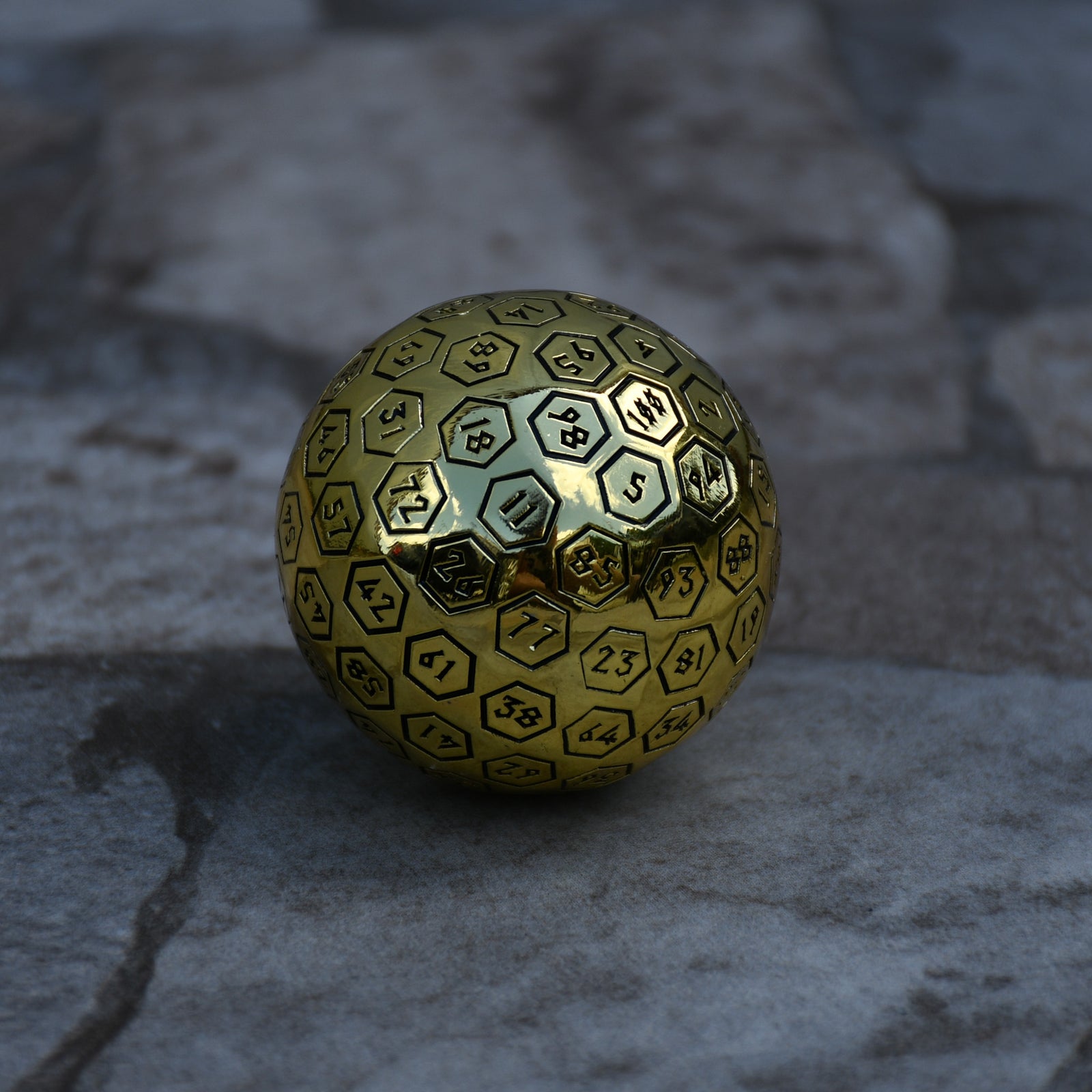 A large gold metal d100 dice with black engraved numbers, sitting on a rocky surface.