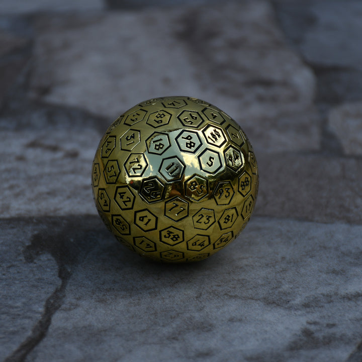 A large gold metal d100 dice with black engraved numbers, sitting on a rocky surface.