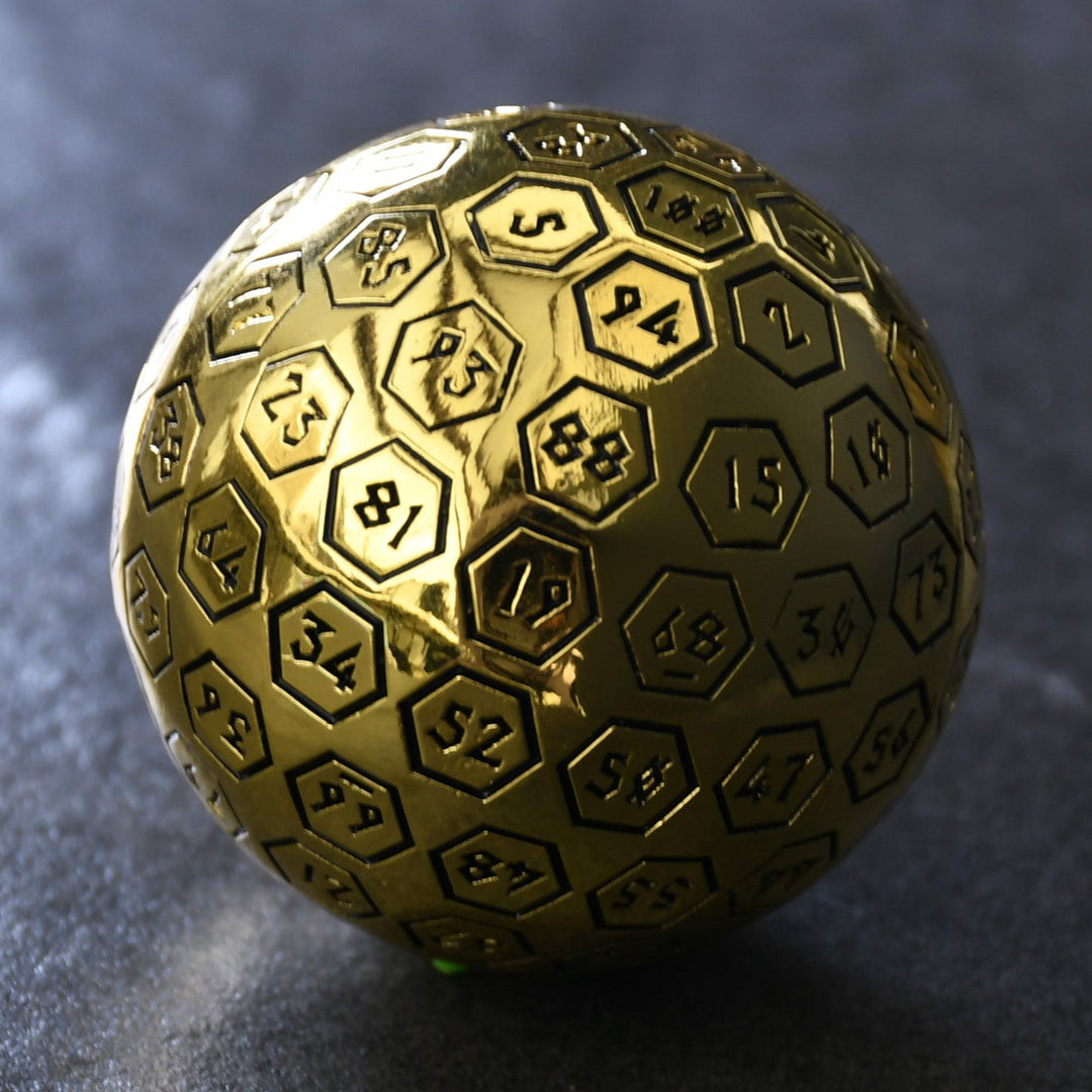 A large gold metal d100 dice with black engraved numbers, sitting on a rocky surface.