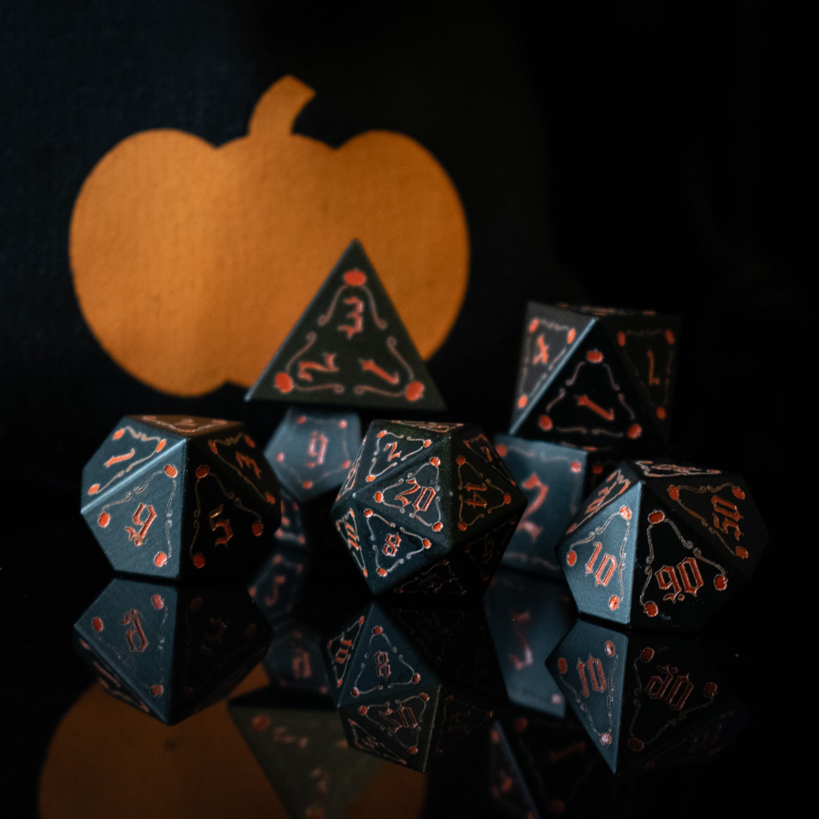 A set of black aluminum metal dice with orange engraved numbers, displayed in front of a pumpkin-shaped silhouette. The dice include a D4, D6, D8, two D10s, D12, and D20.