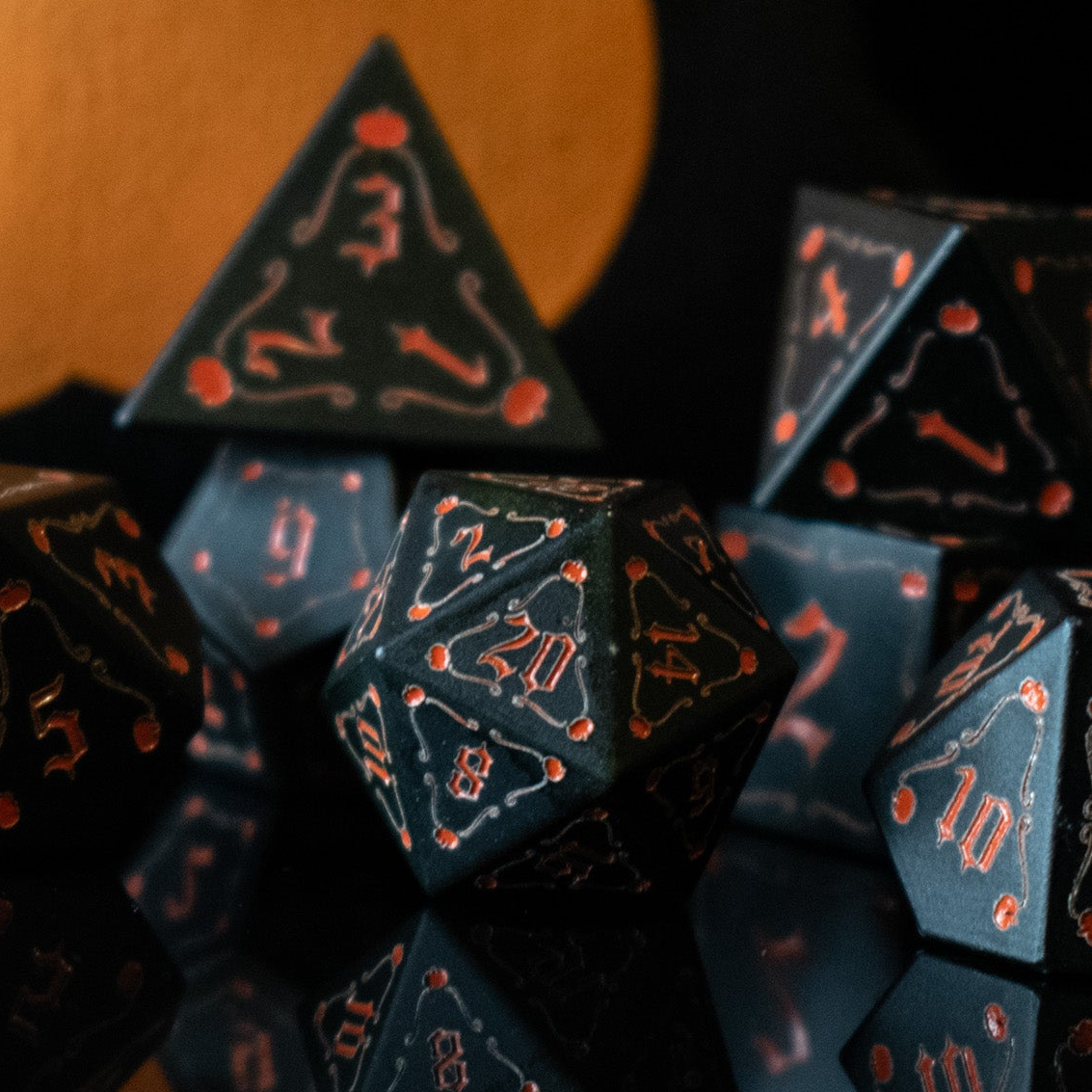 A set of black aluminum metal dice with orange engraved numbers, displayed in front of a pumpkin-shaped silhouette. The dice include a D4, D6, D8, two D10s, D12, and D20.