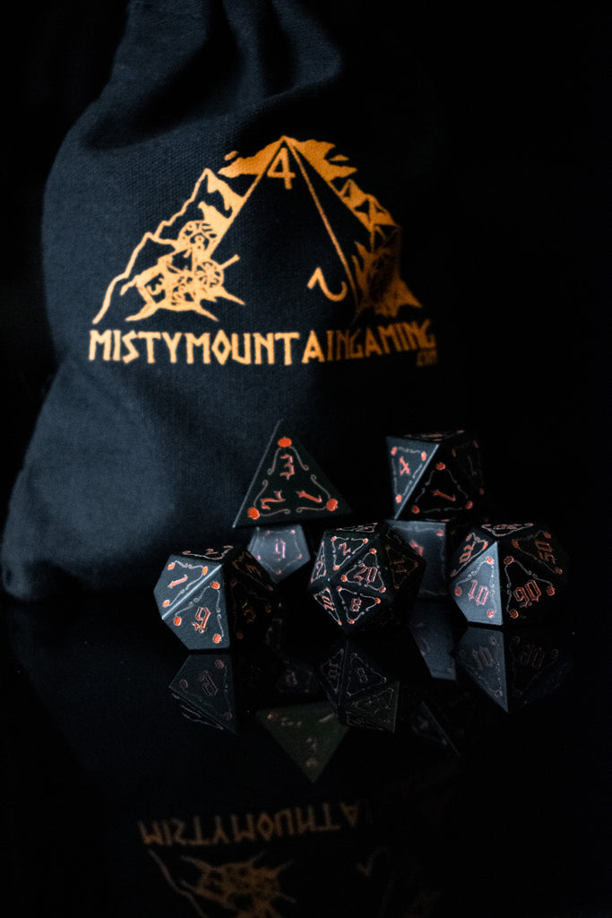 A set of black aluminum metal dice with orange engraved numbers, displayed in front of a pumpkin-shaped silhouette. The dice include a D4, D6, D8, two D10s, D12, and D20.