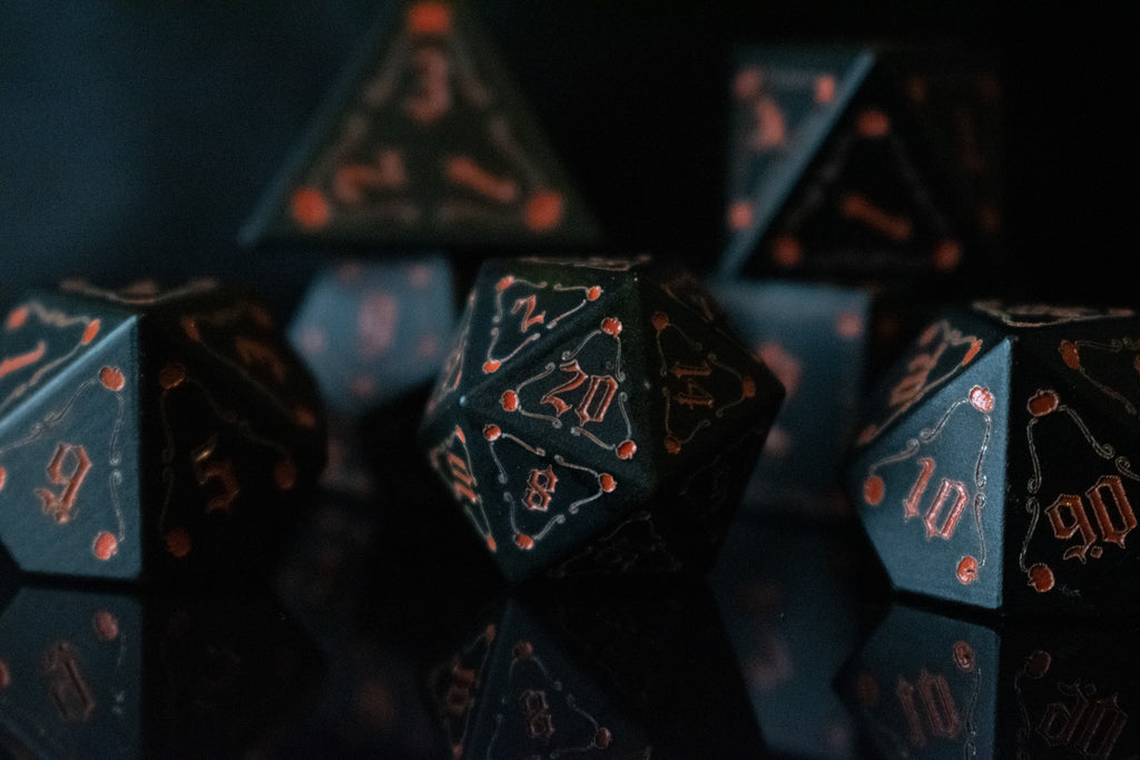 A set of black aluminum metal dice with orange engraved numbers, displayed in front of a pumpkin-shaped silhouette. The dice include a D4, D6, D8, two D10s, D12, and D20.