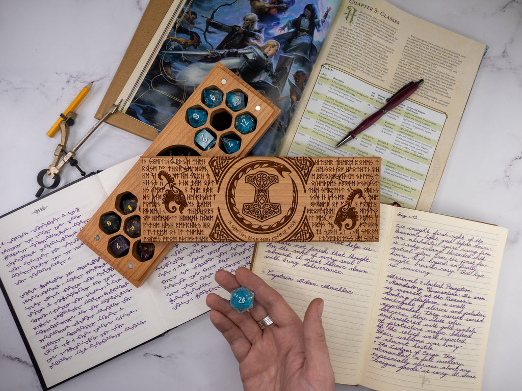 Wooden dice tray with engraved symbols on a marble surface with books and a pen.