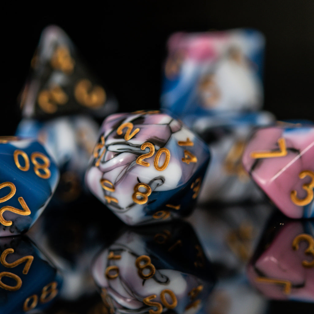 A set of acrylic dice with engraved numbers, featuring a variety of colors including blue, pink, white, and black, displayed on a reflective surface for playing tabletop games war games and board games