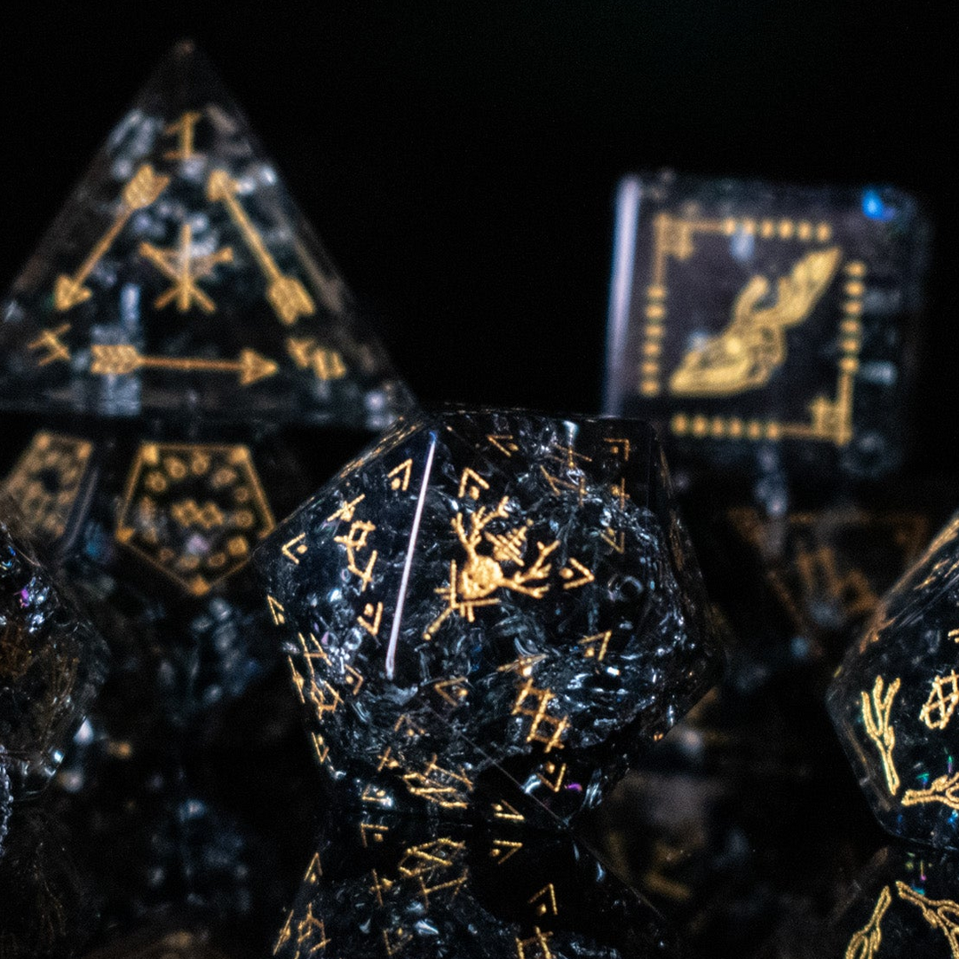 A collection of hand-carved, smoky-gray glass dice with gold engraved numbers and wendigo inspired details, displayed against a black background.