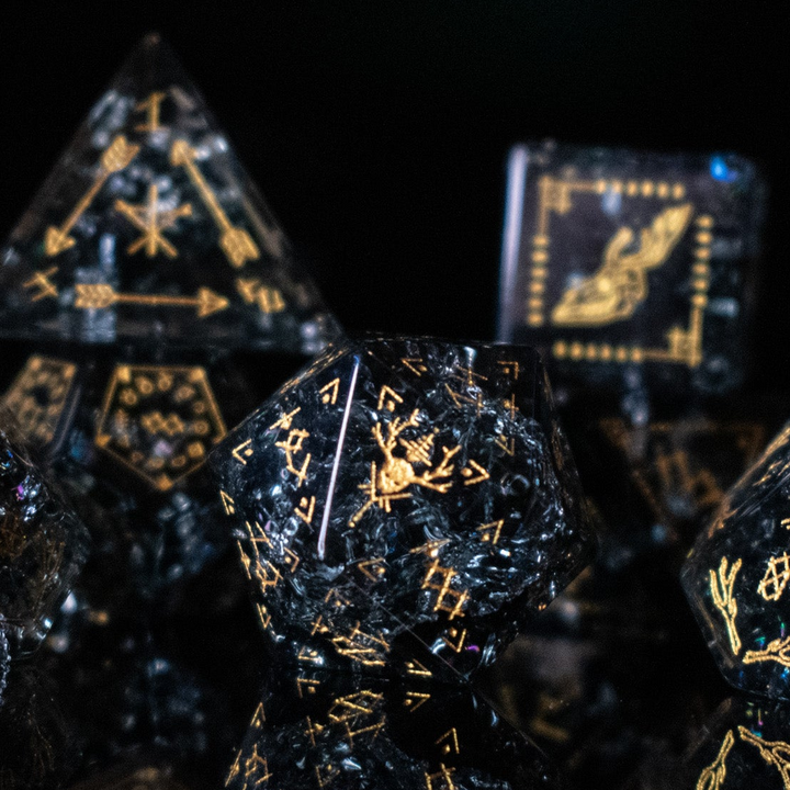 A collection of hand-carved, smoky-gray glass dice with gold engraved numbers and wendigo inspired details, displayed against a black background.