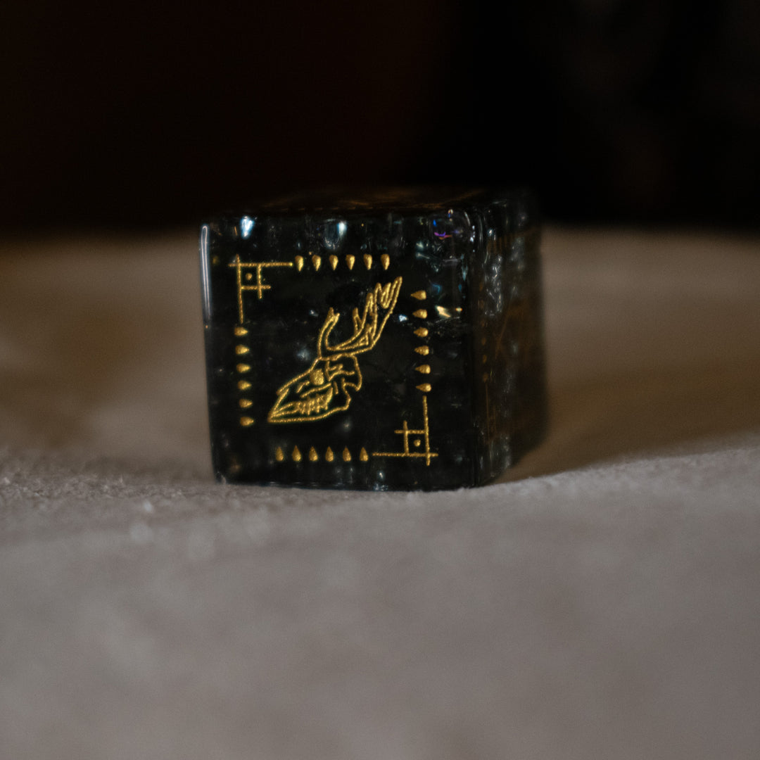 hand-carved, smoky-gray glass dice with gold engraved numbers and wendigo inspired details, displayed against a black background.